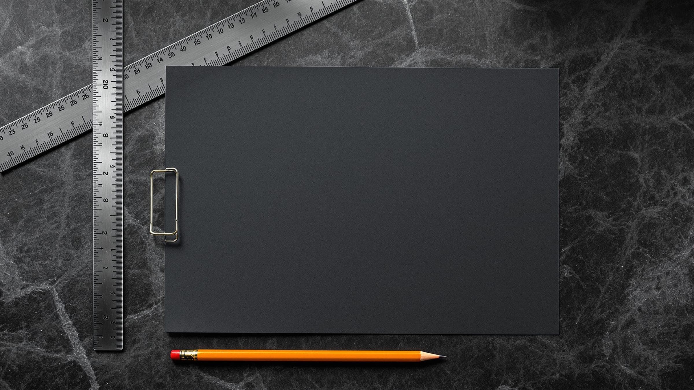 A clipboard of blank dark paper under a steel ruler, an orange pencil lying below it.