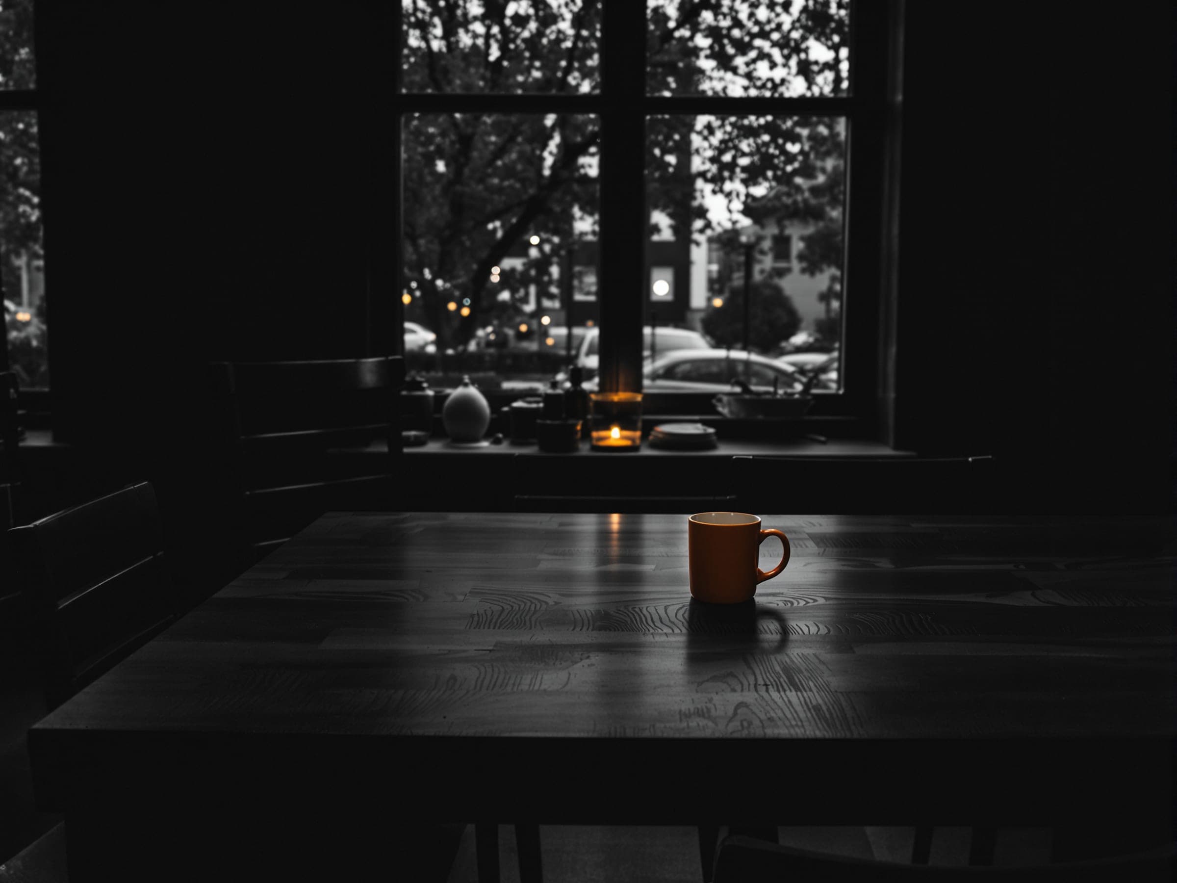 An empty dark dining table by a window in the evening, a single orange mug on it.