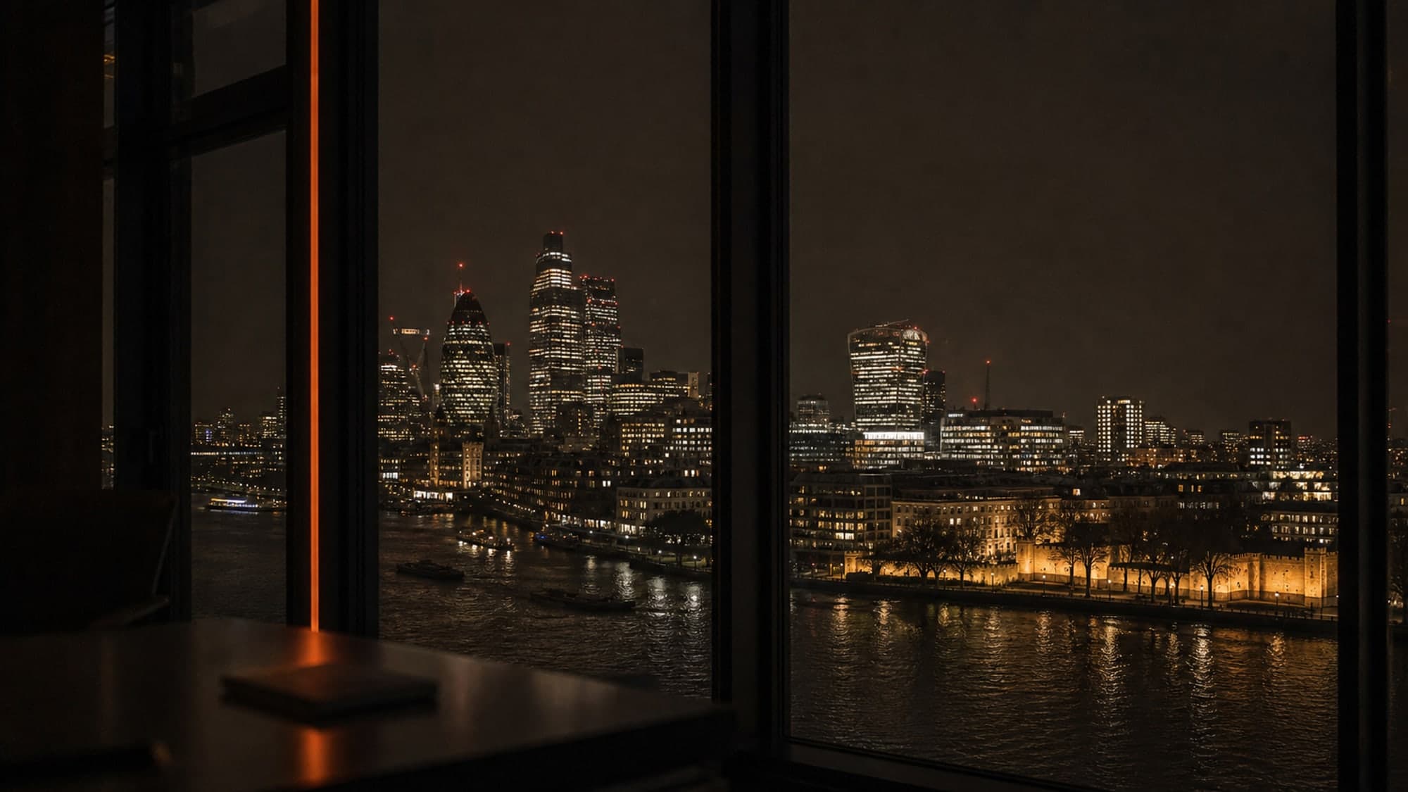 The London skyline at night through a tall office window, the City towers and the river lit below.