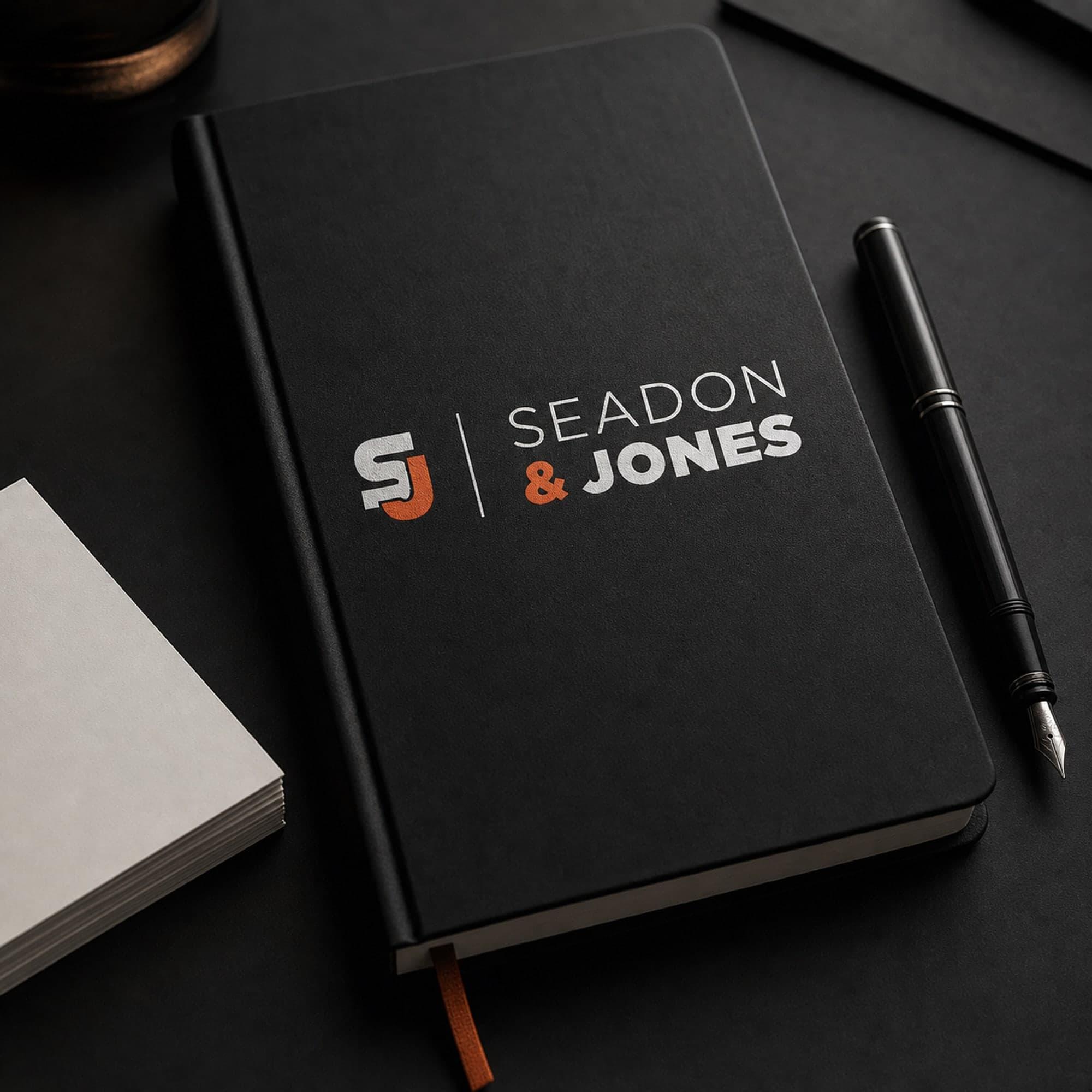 A black notebook with the Seadon & Jones logo on its cover, a fountain pen and blank index cards on a dark desk.