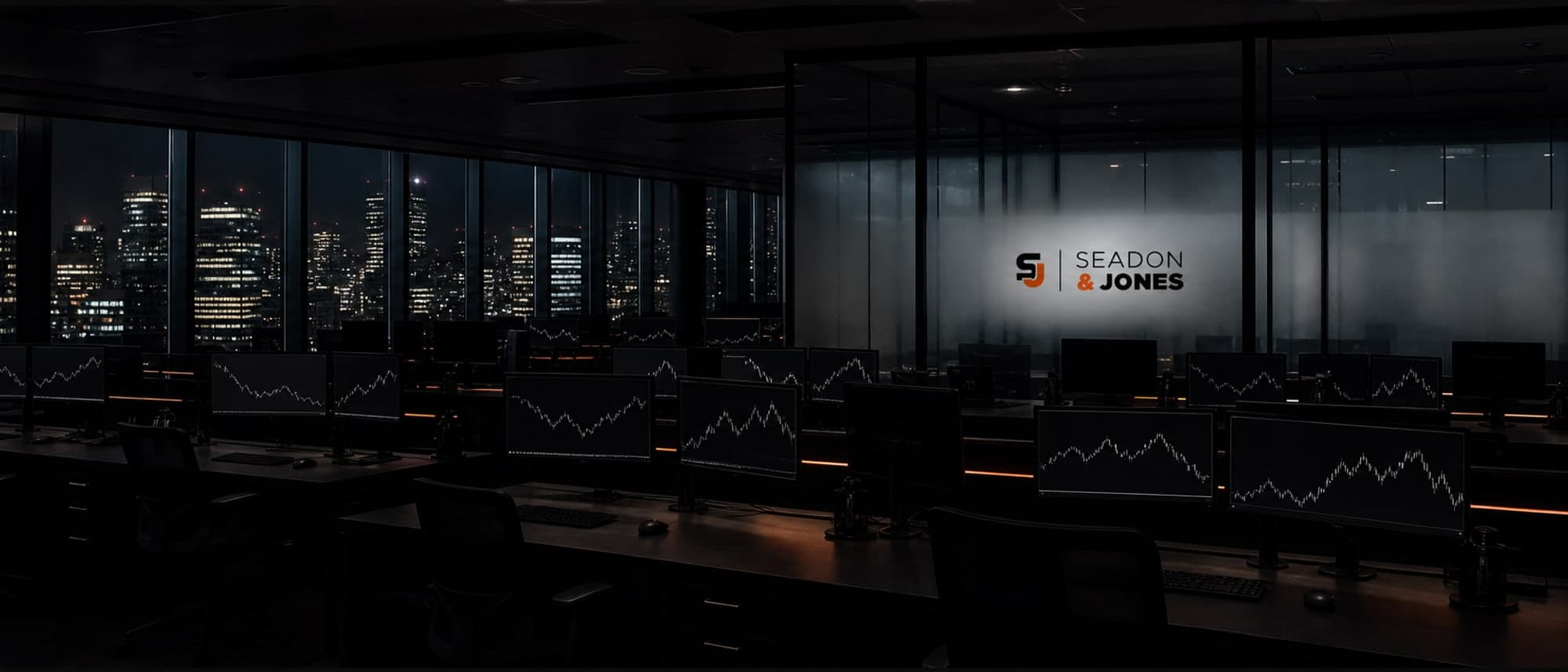 An empty open plan trading office after hours, screens of grey line charts and the Seadon & Jones logo on glass.