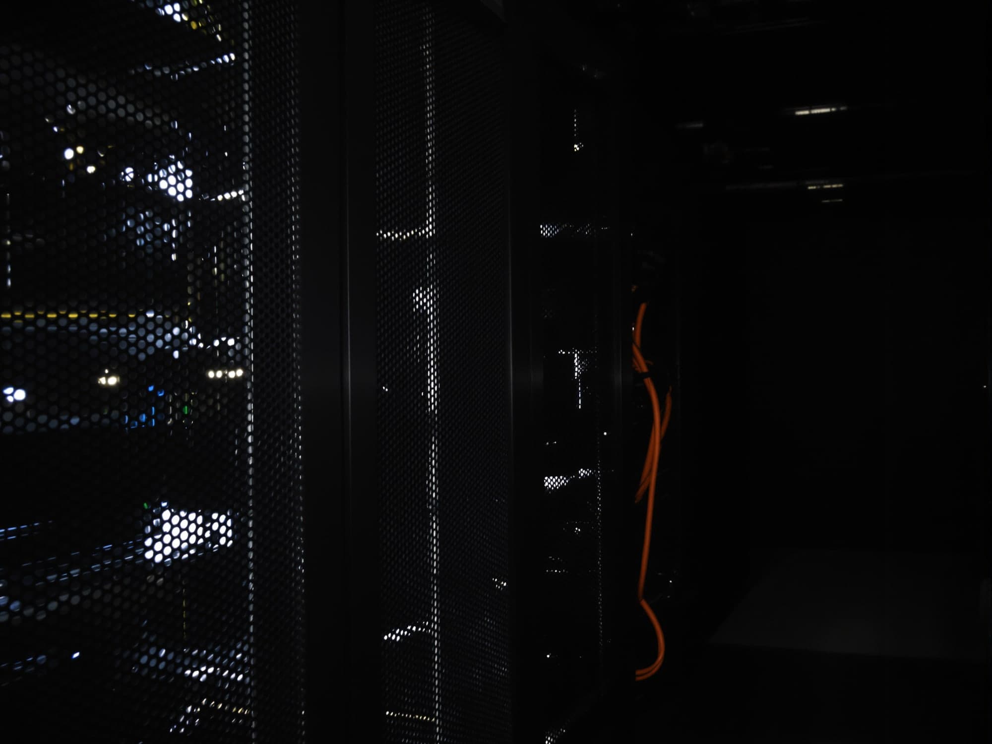 Dark server racks behind perforated doors in a dim room, one orange cable among the black ones.