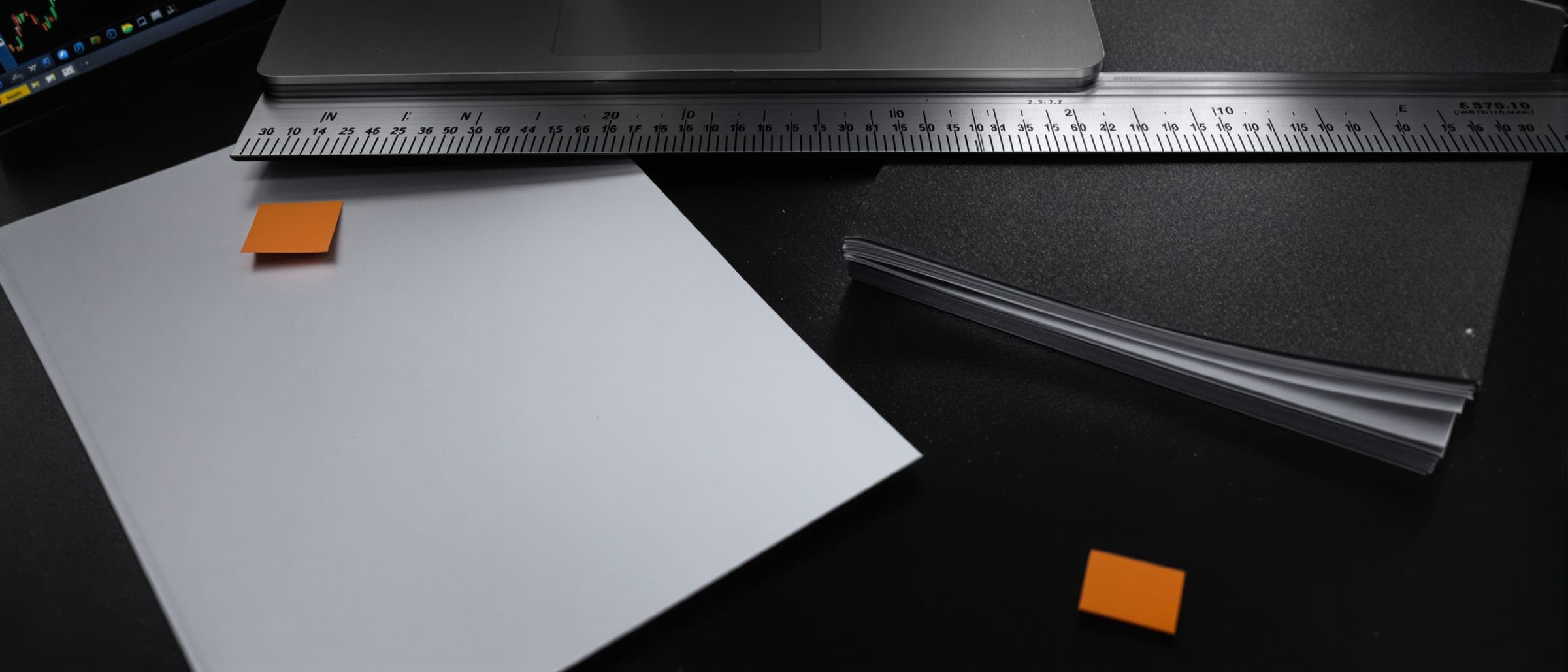Blank pages and a steel ruler on a dark desk, small orange tabs beside them.