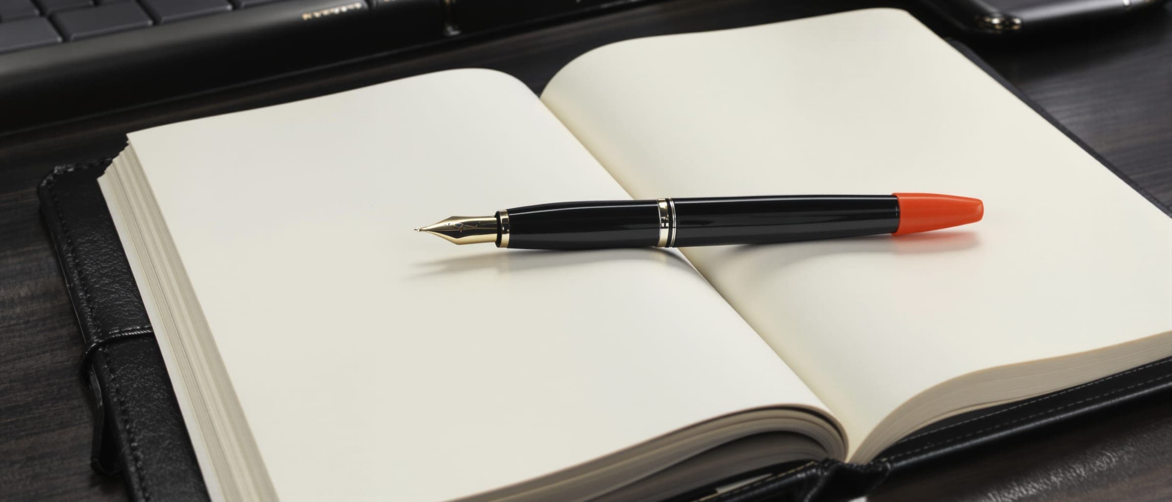 A notebook lying open on its blank pages, a black fountain pen with an orange cap across it.