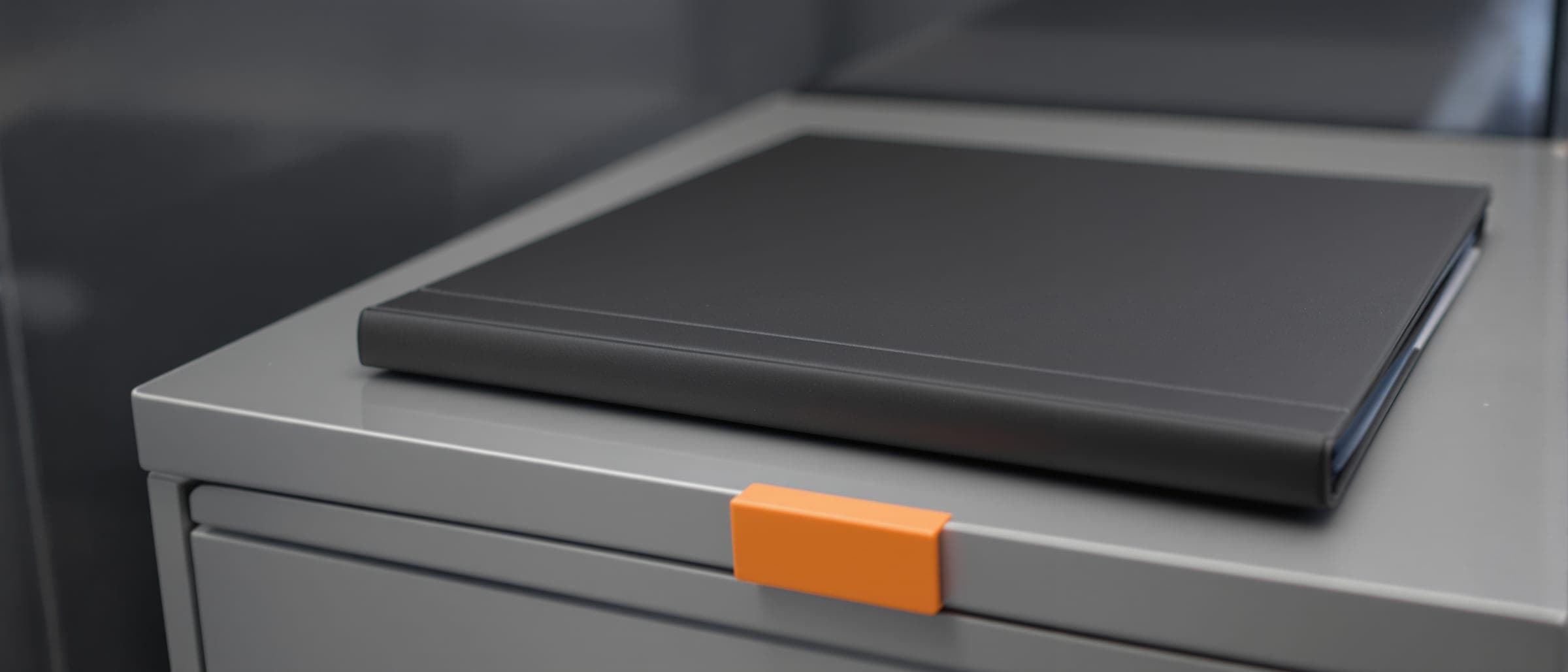 A closed dark folder on a steel filing drawer with an orange tab.