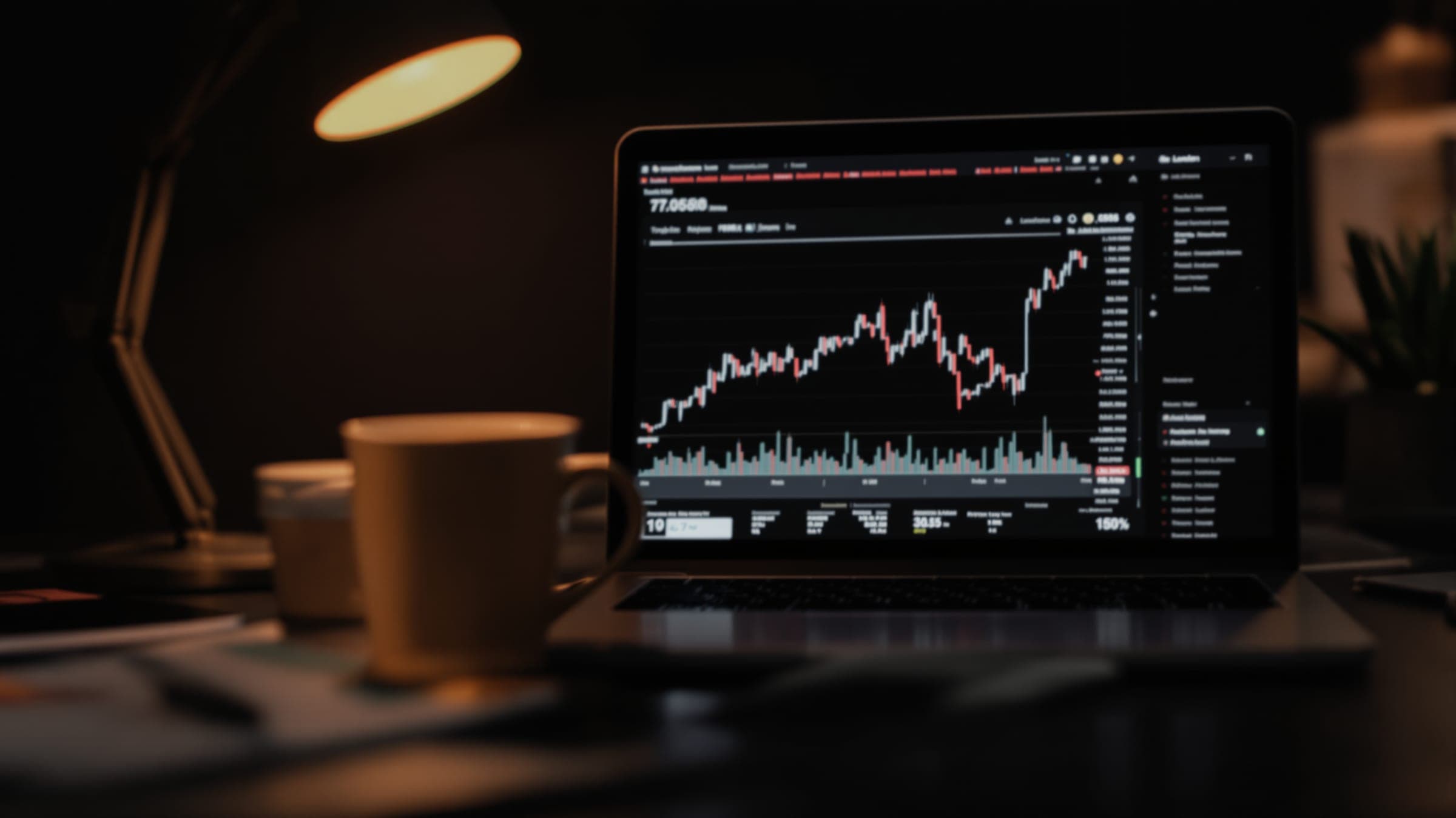 A laptop showing a price chart on a dark desk, a cup beside it under a warm lamp.