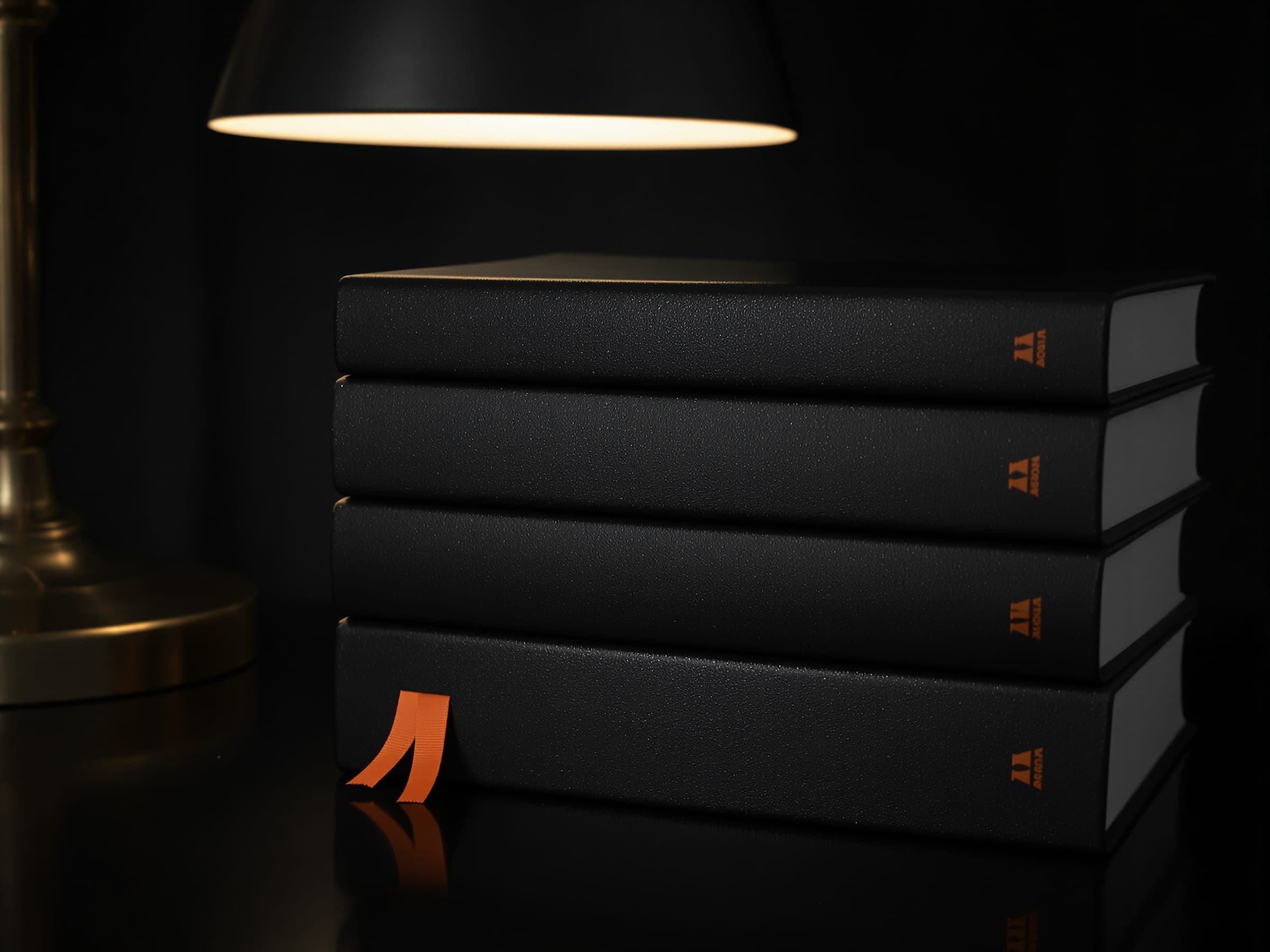 A stack of dark hardback books under a desk lamp, an orange ribbon marker beside them.