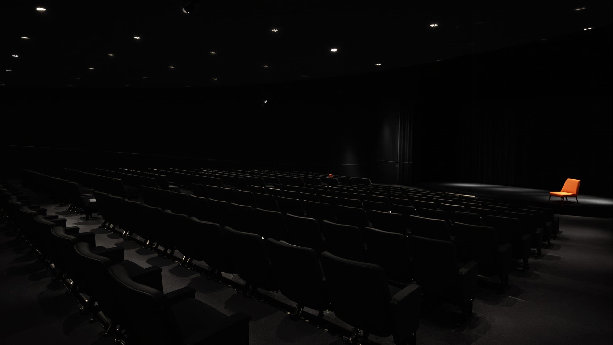 Rows of empty seats in a dark theatre, one orange chair lit on the stage.