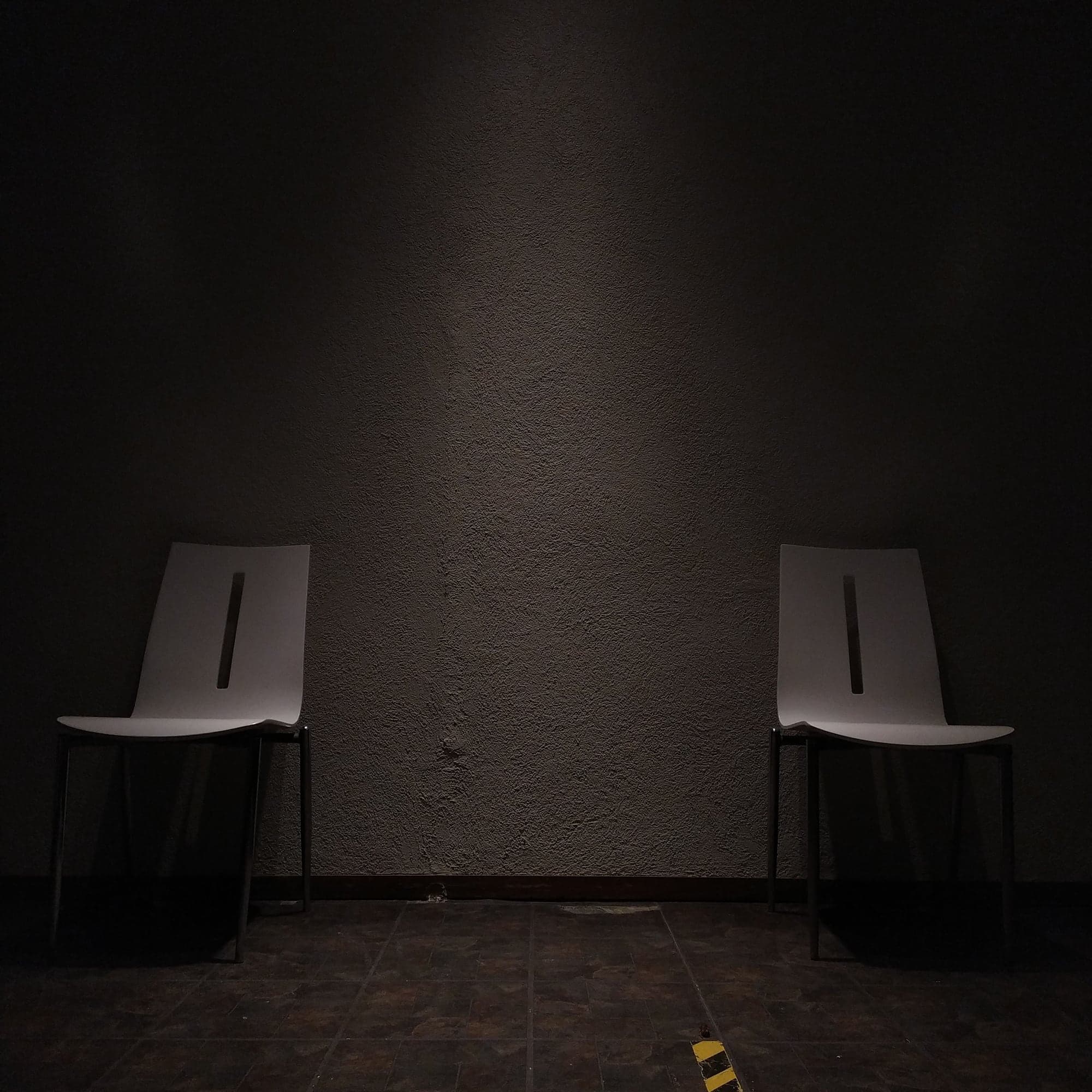 Two empty white chairs against a dark textured wall, a soft light falling between them.