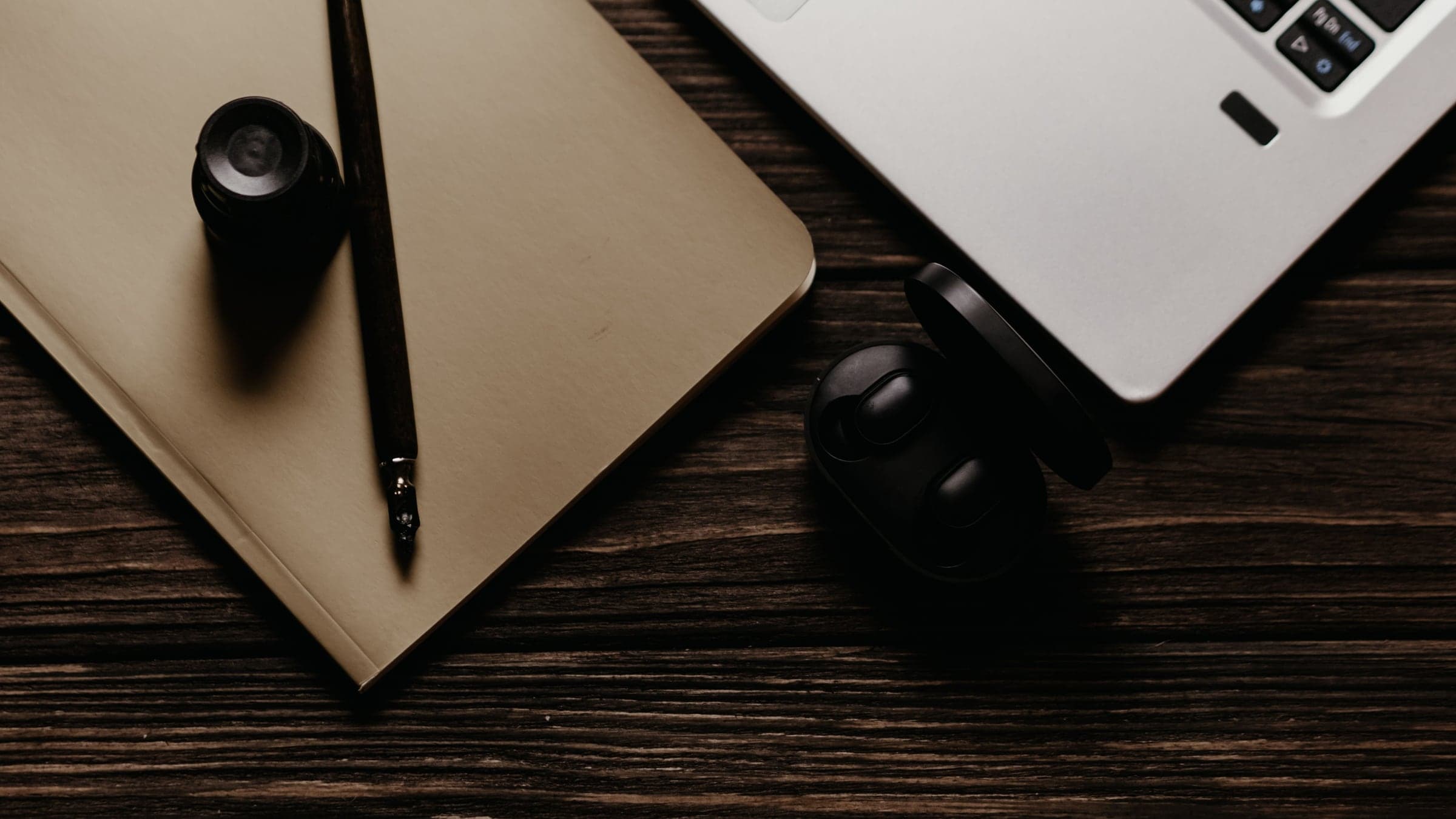 A notebook, a pen and a laptop on a dark wooden desk, seen from above.
