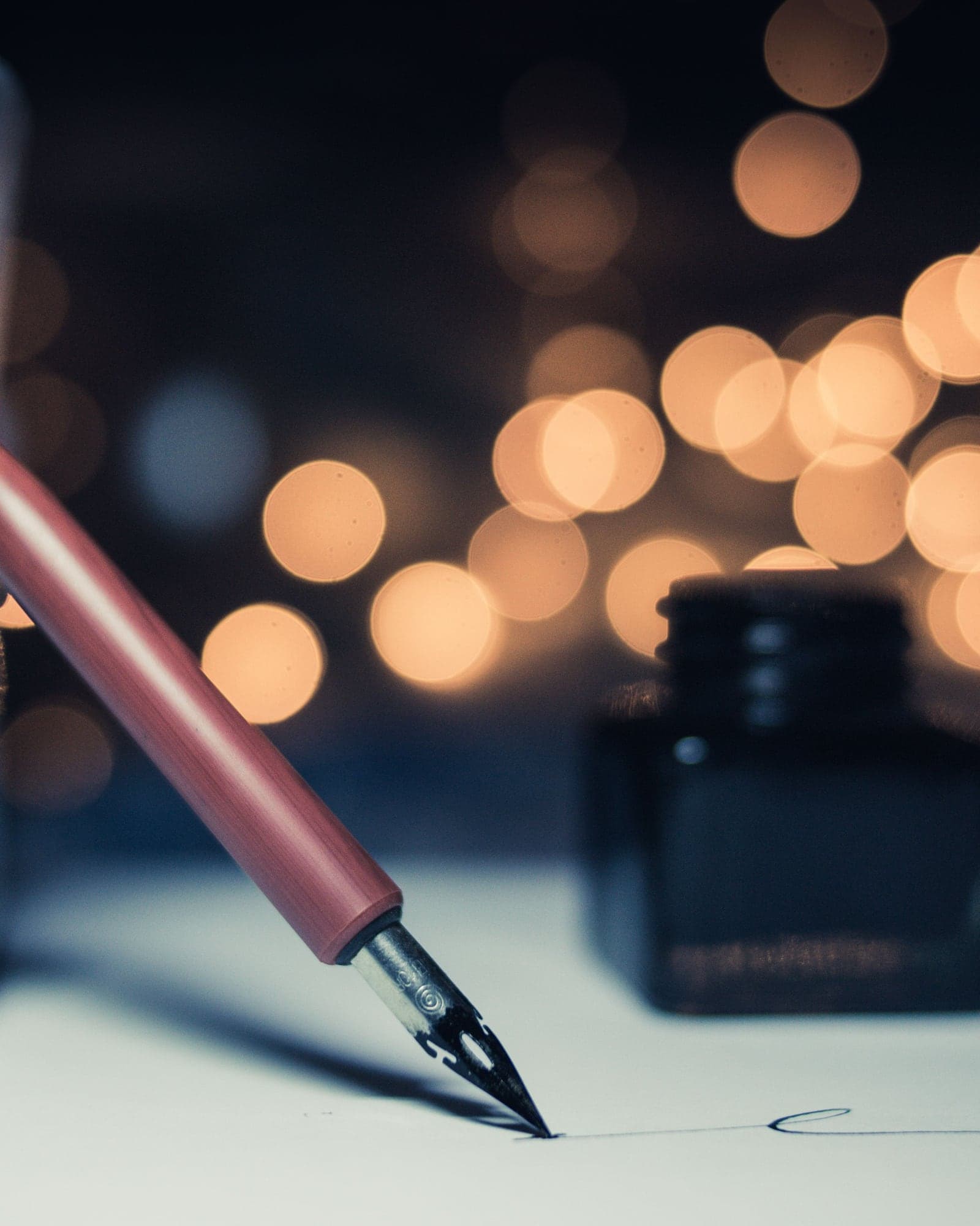 A fountain pen writing on paper beside an ink bottle, warm lights glowing out of focus behind.