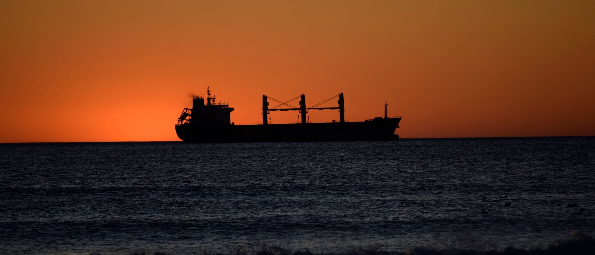 A cargo ship silhouetted on the horizon against an orange sky at dusk.