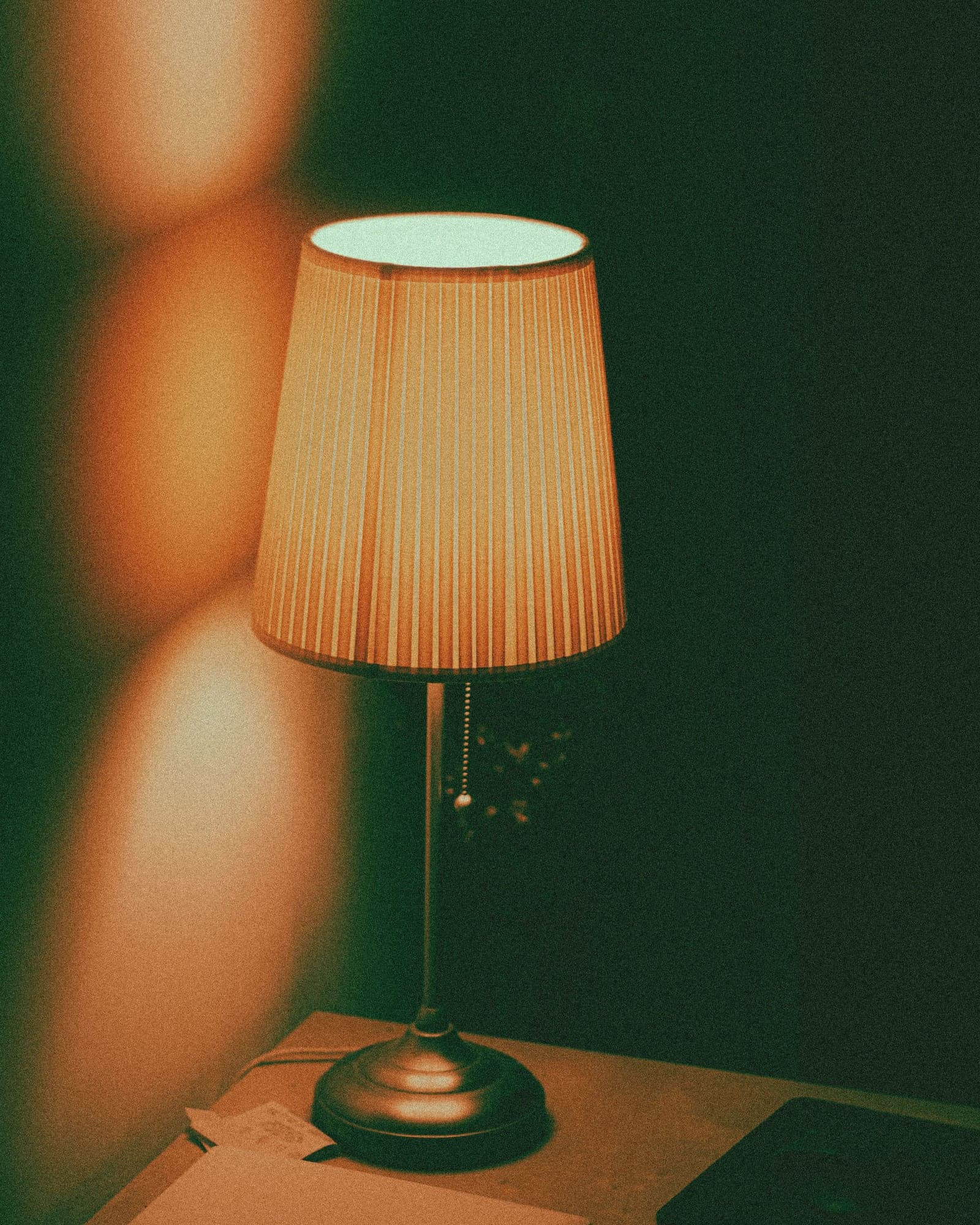 A table lamp with a pleated shade glowing on a desk against a dark green wall.