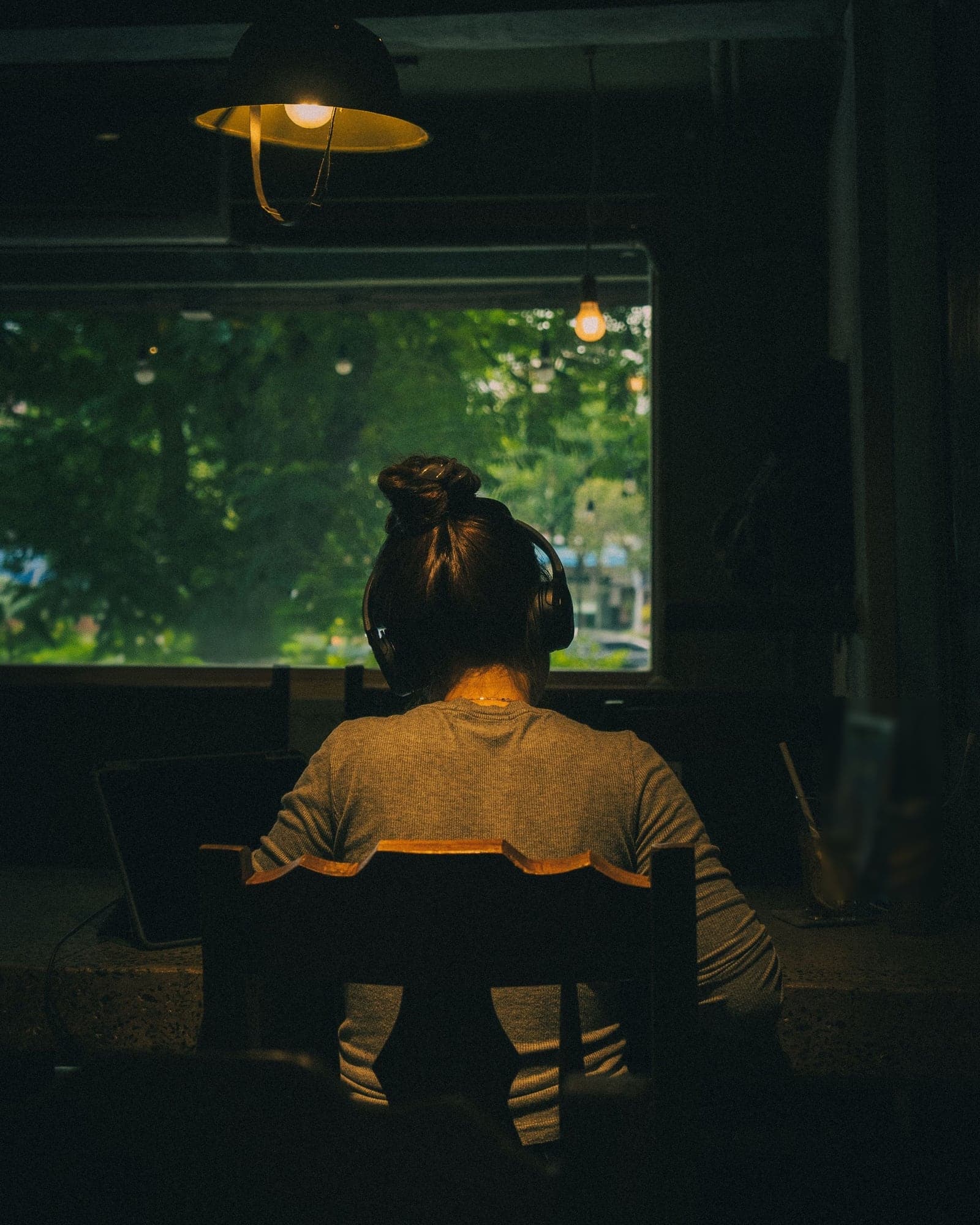 A young person with headphones studying at a desk under a hanging lamp, seen from behind.