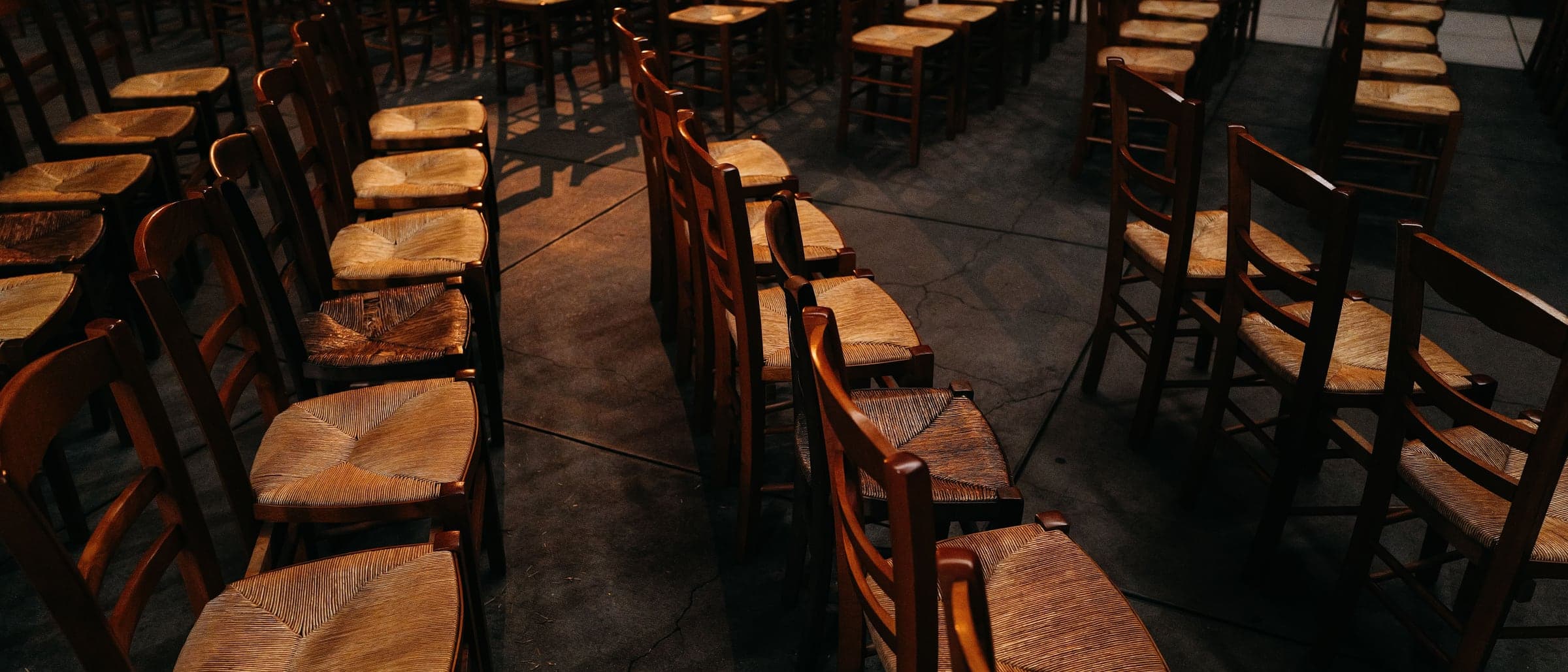 Rows of empty wooden chairs with woven seats set out in a dim hall.