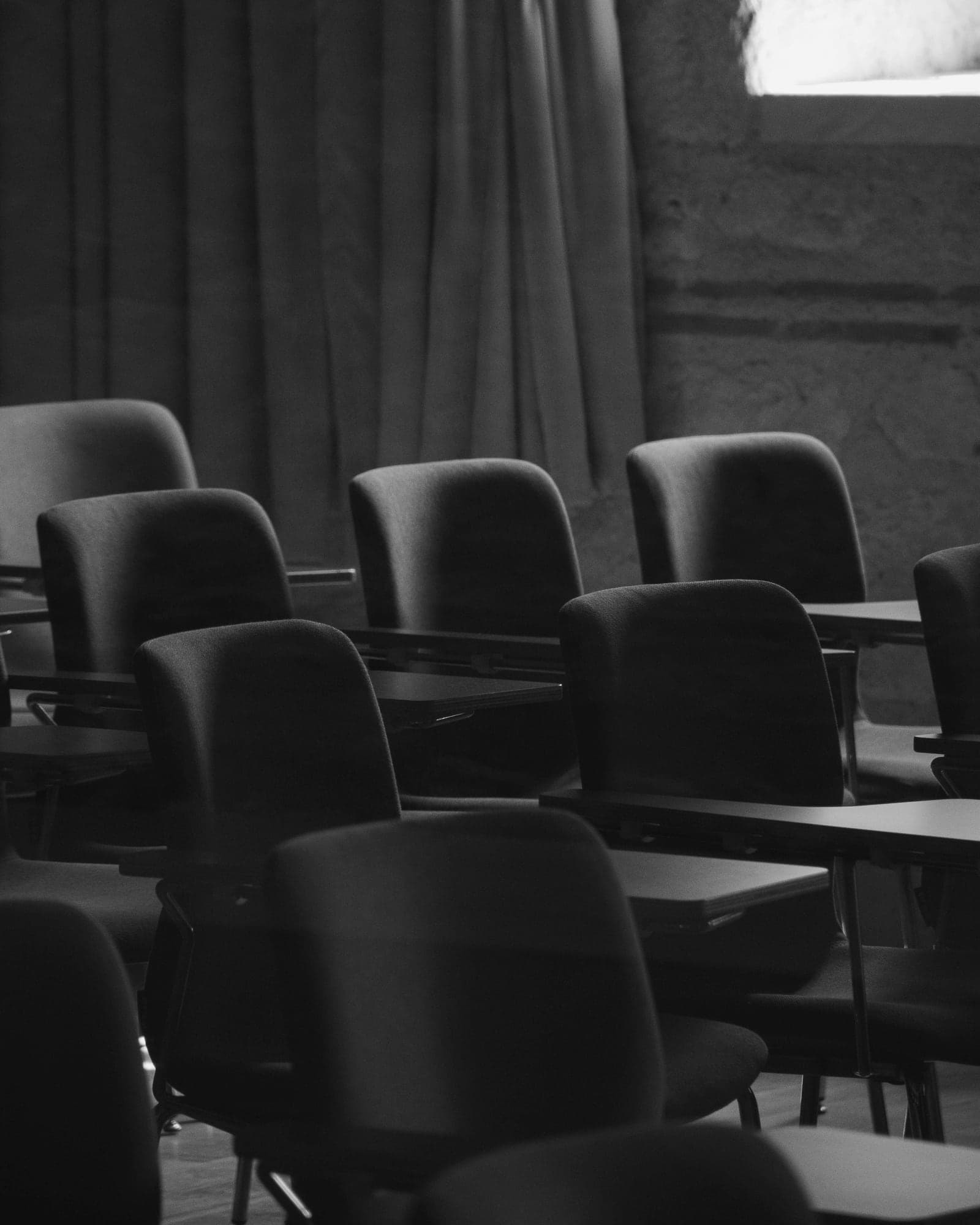 A dim, empty classroom with rows of dark chairs and tables beside a heavy curtain.