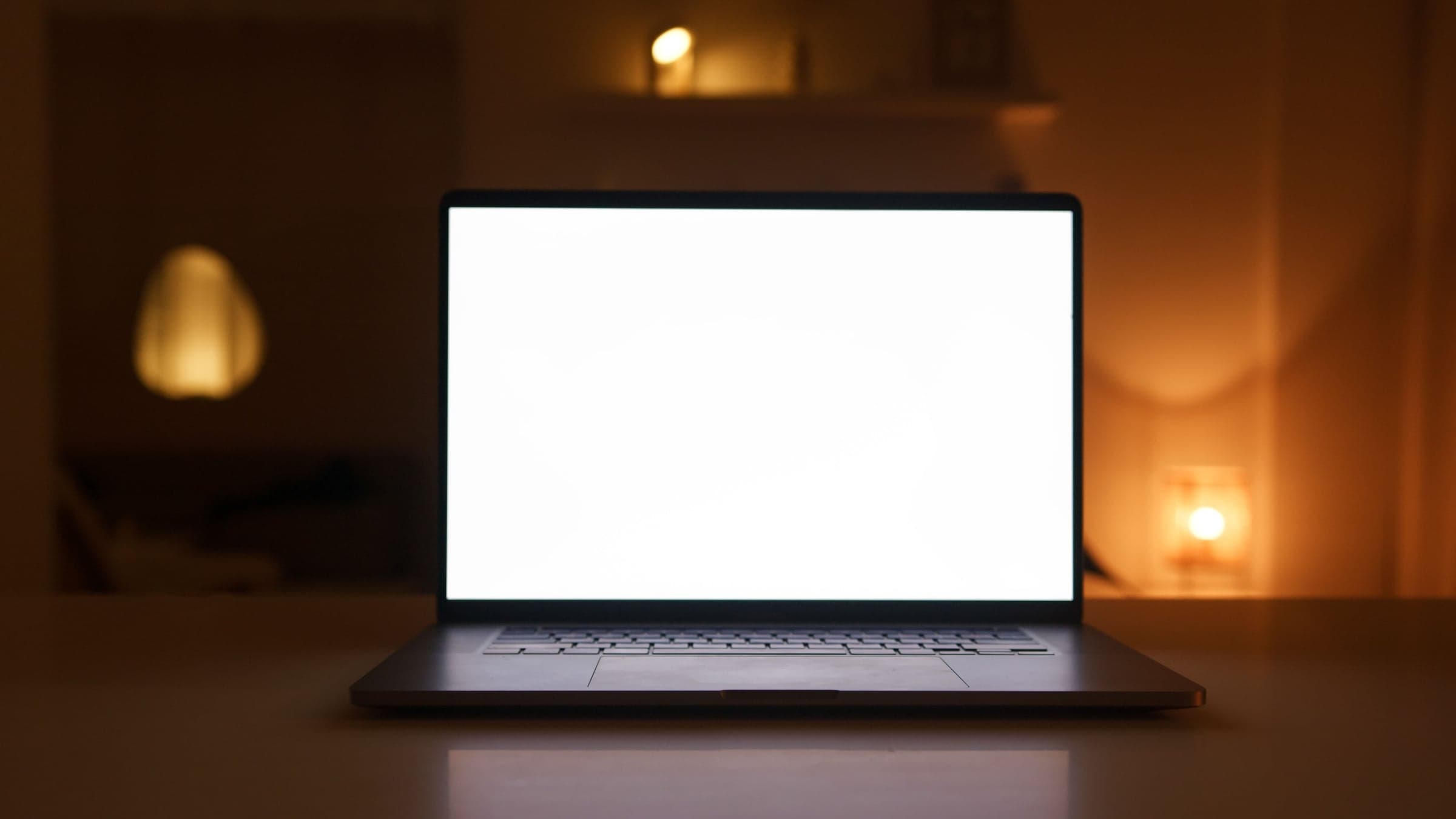 An open laptop with a glowing screen on a desk in a dim room lit by warm lamps.