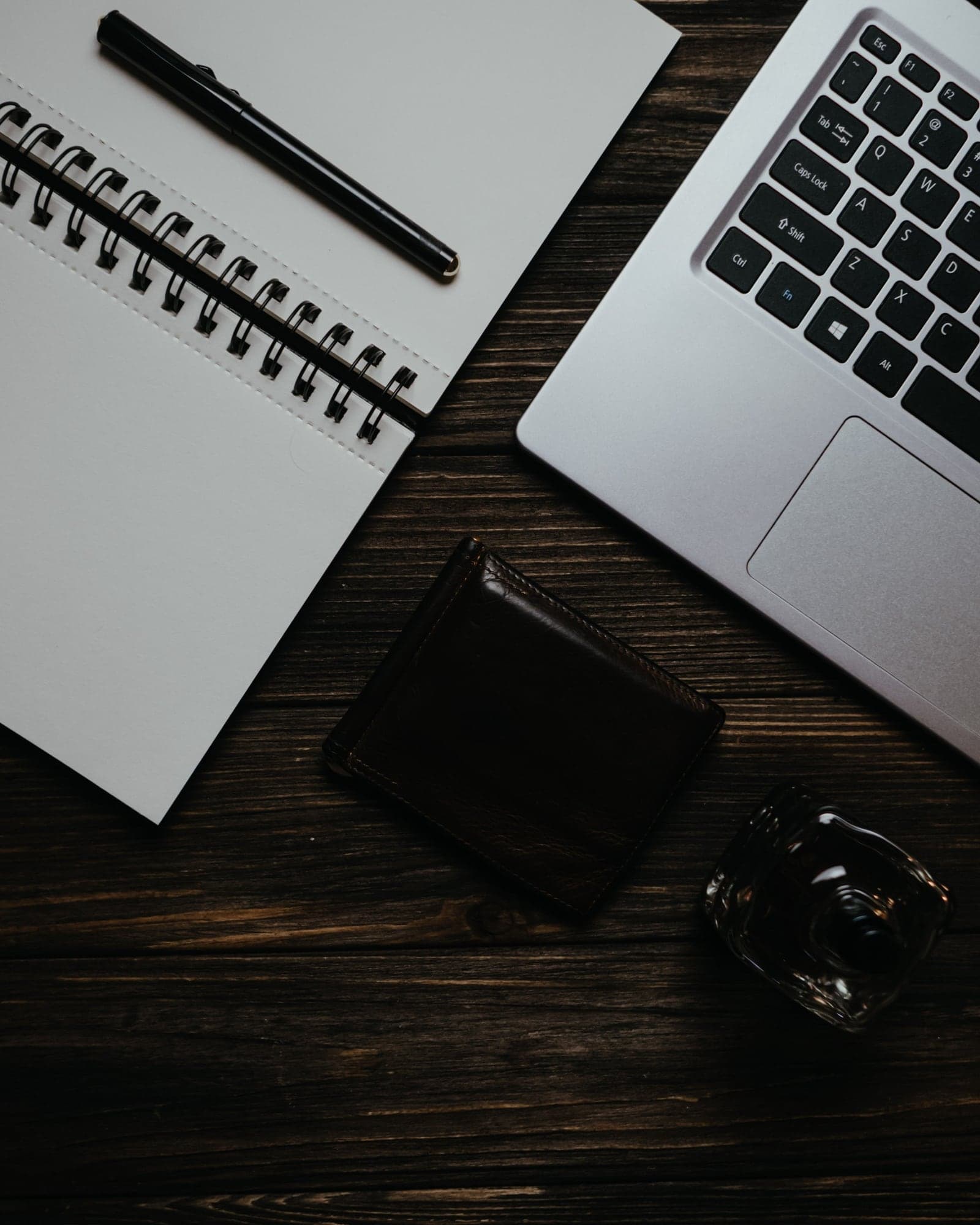 An open notebook, a pen, a laptop and a wallet laid out on a dark wooden desk.