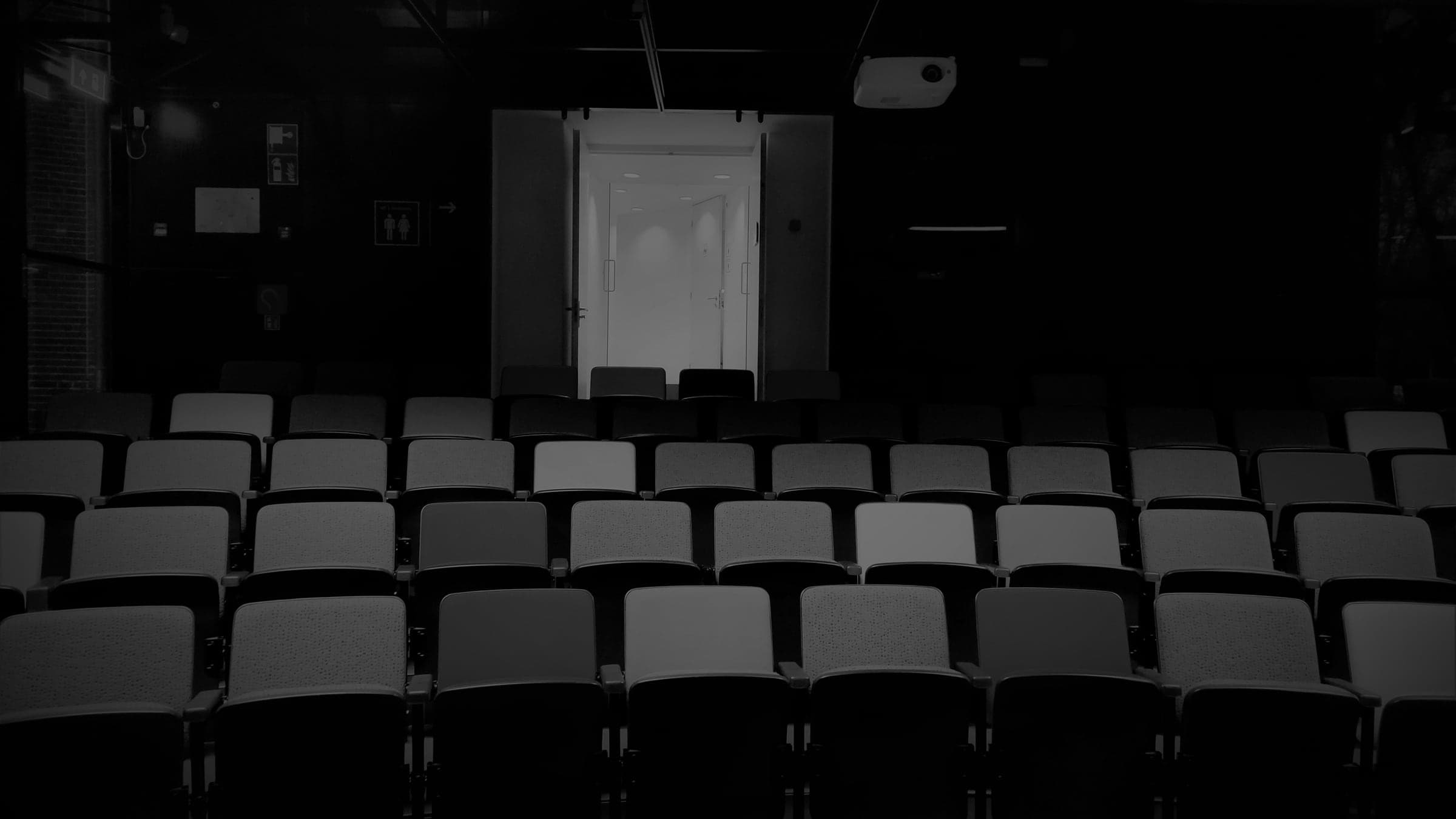 Rows of empty seats in a dark auditorium facing a lit doorway.