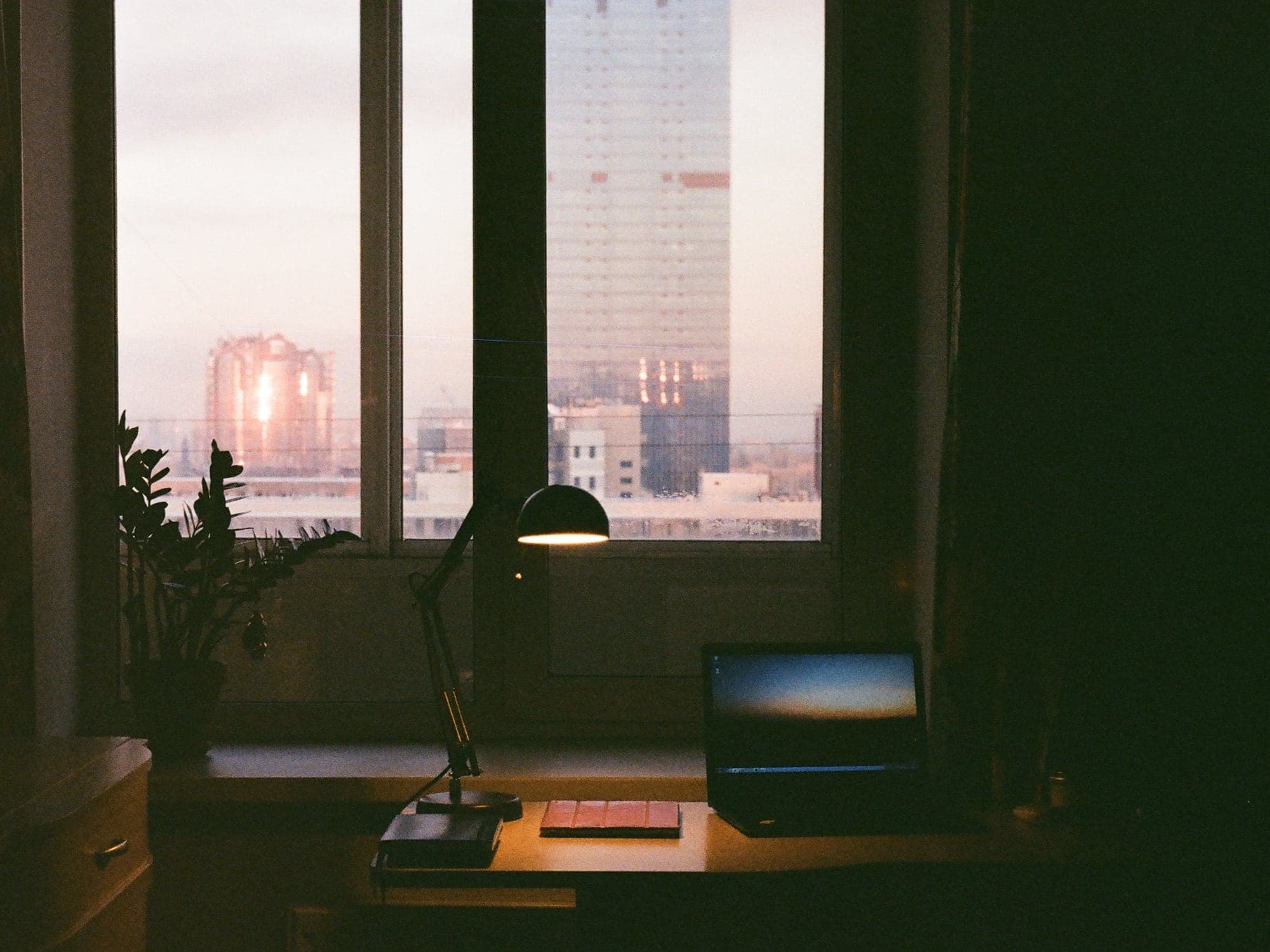A desk with a lamp and a laptop by a window looking out over city towers at dusk.
