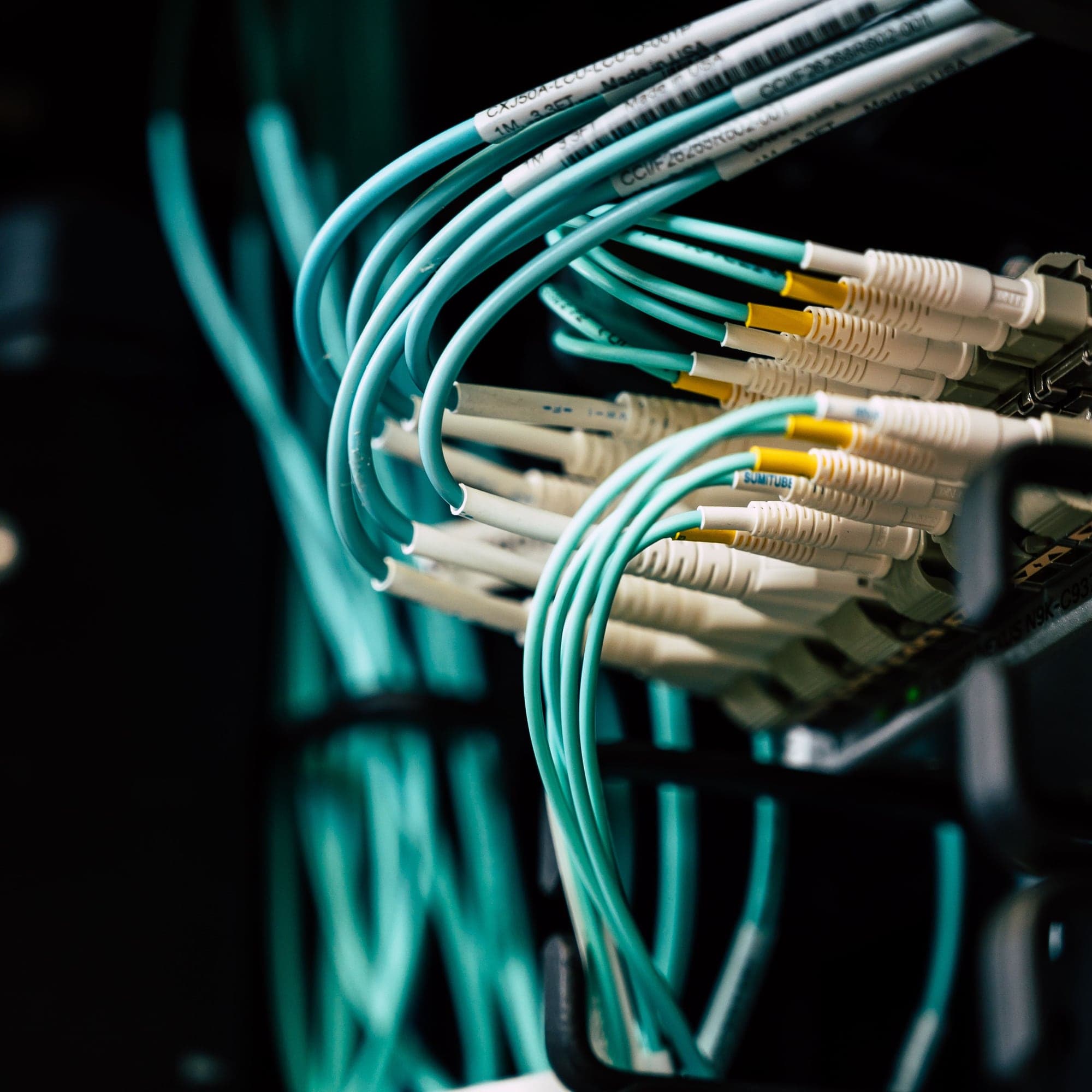 Teal fibre optic cables plugged into rows of connectors in a dark server rack.