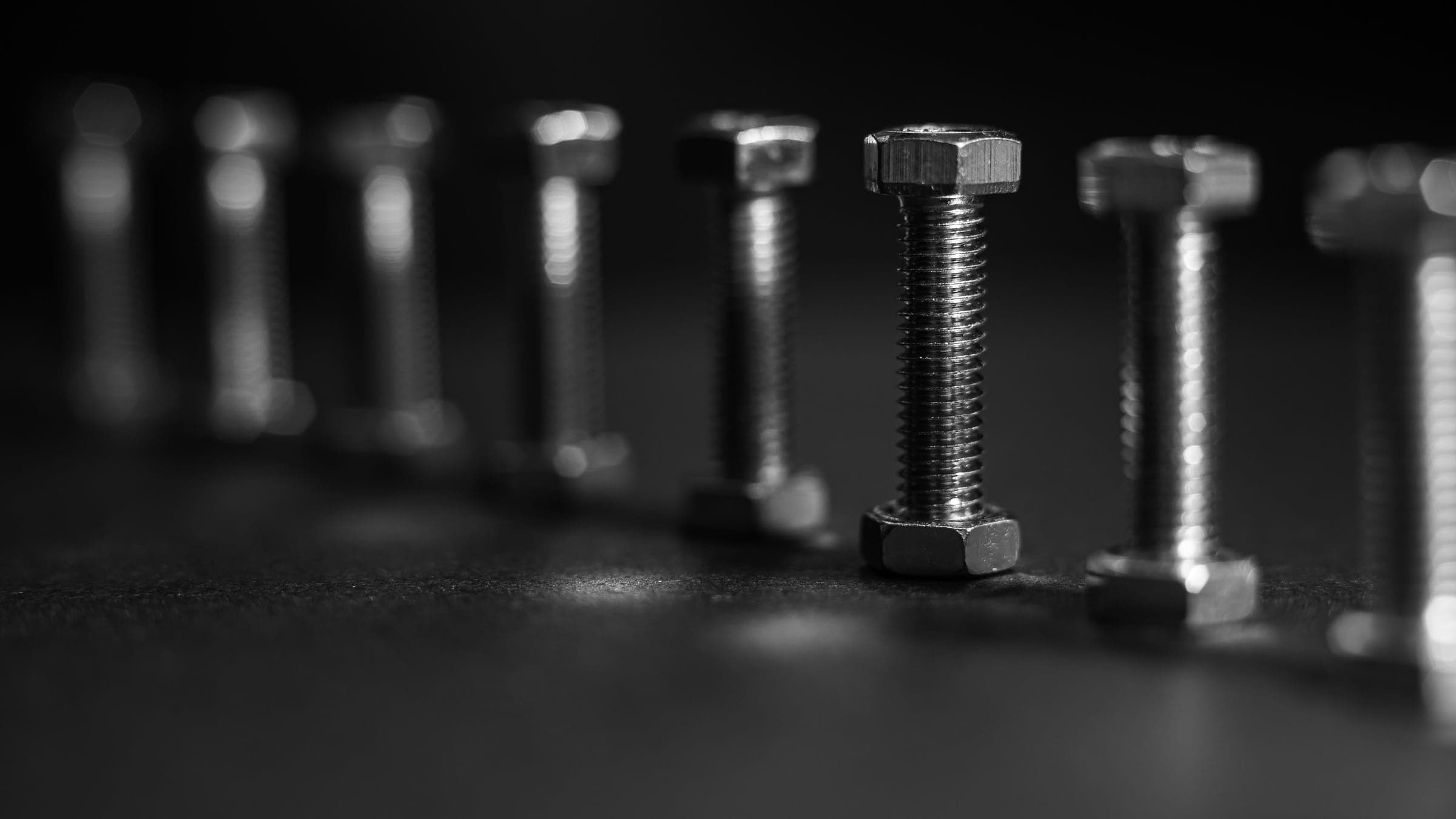 A row of identical steel bolts standing on a dark surface, fading out of focus.