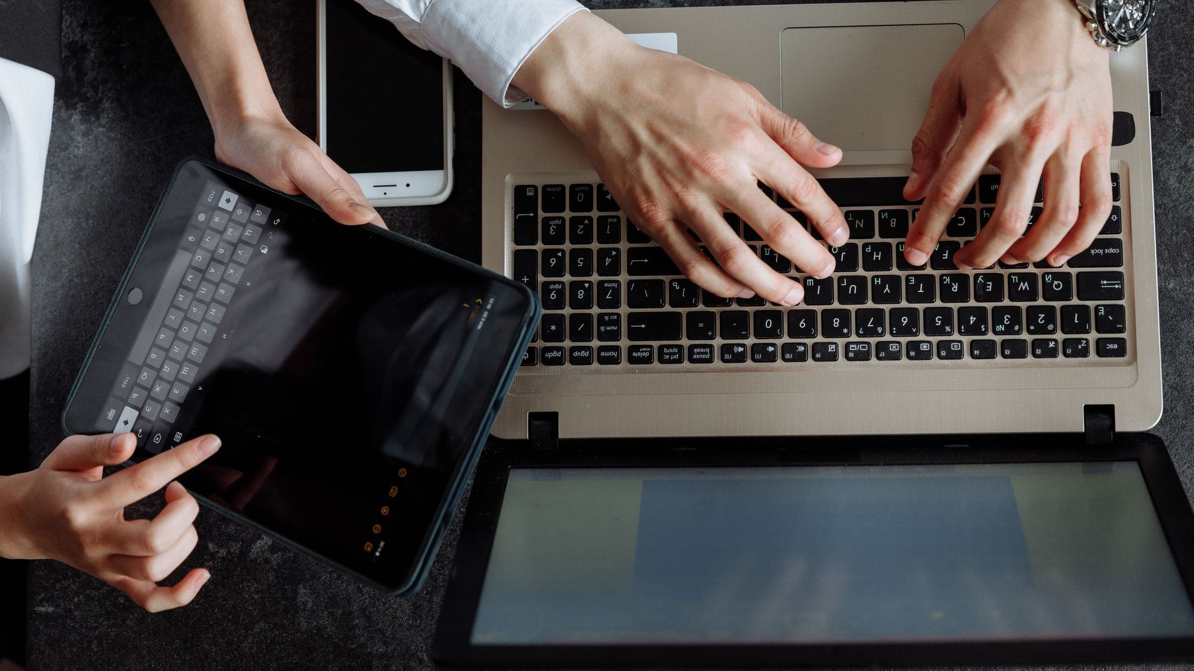 Two people’s hands at work on a laptop and a tablet on a dark desk, seen from above.