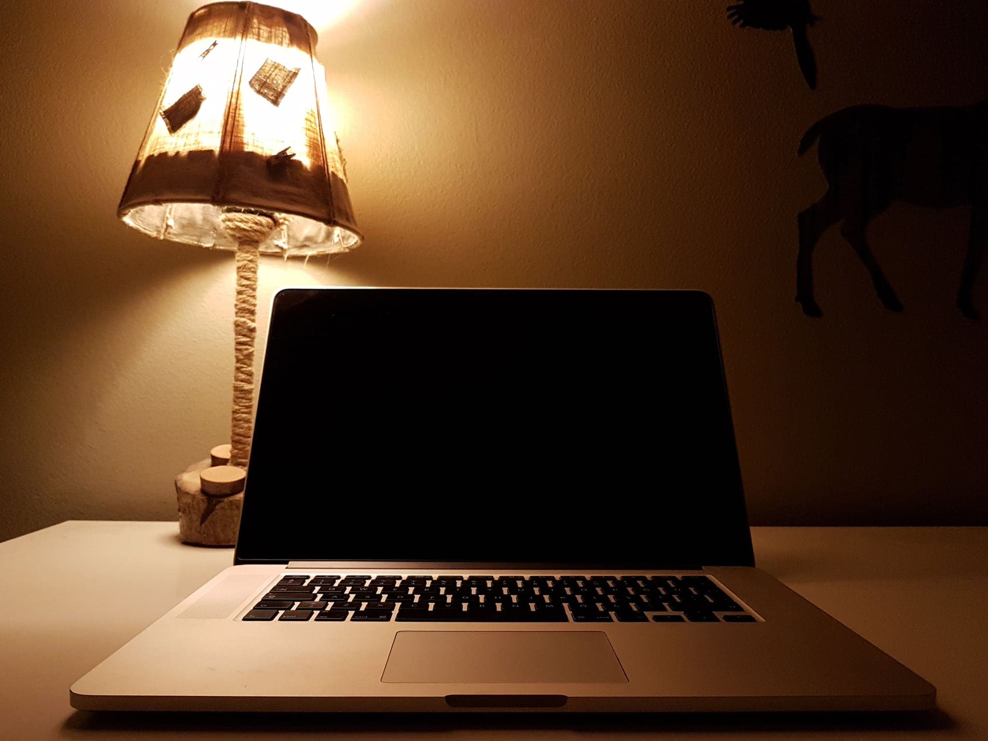 A laptop with a dark screen on a desk beside a warm table lamp at night.