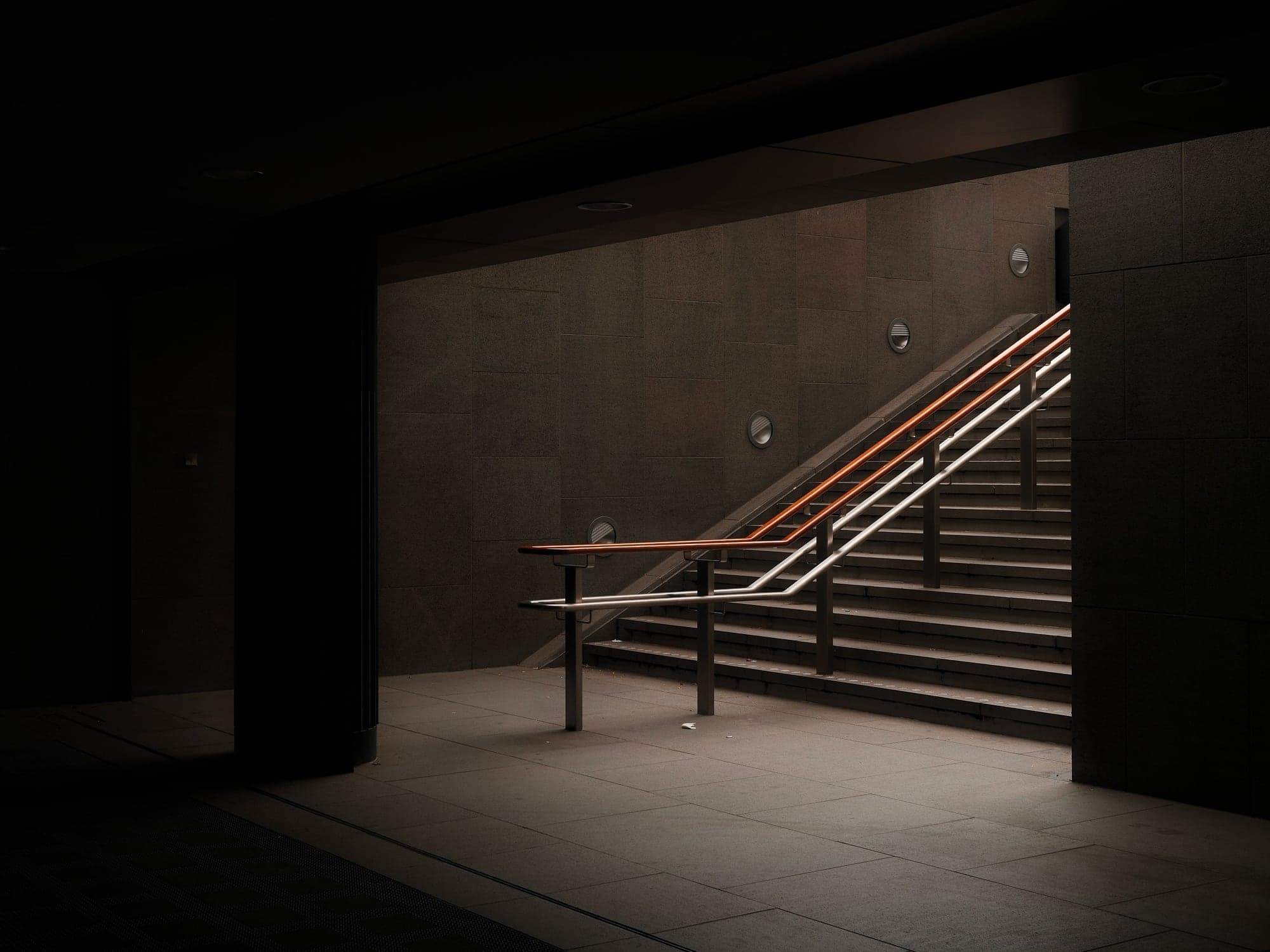 A dark stairwell with a warm lit handrail rising beside concrete steps.