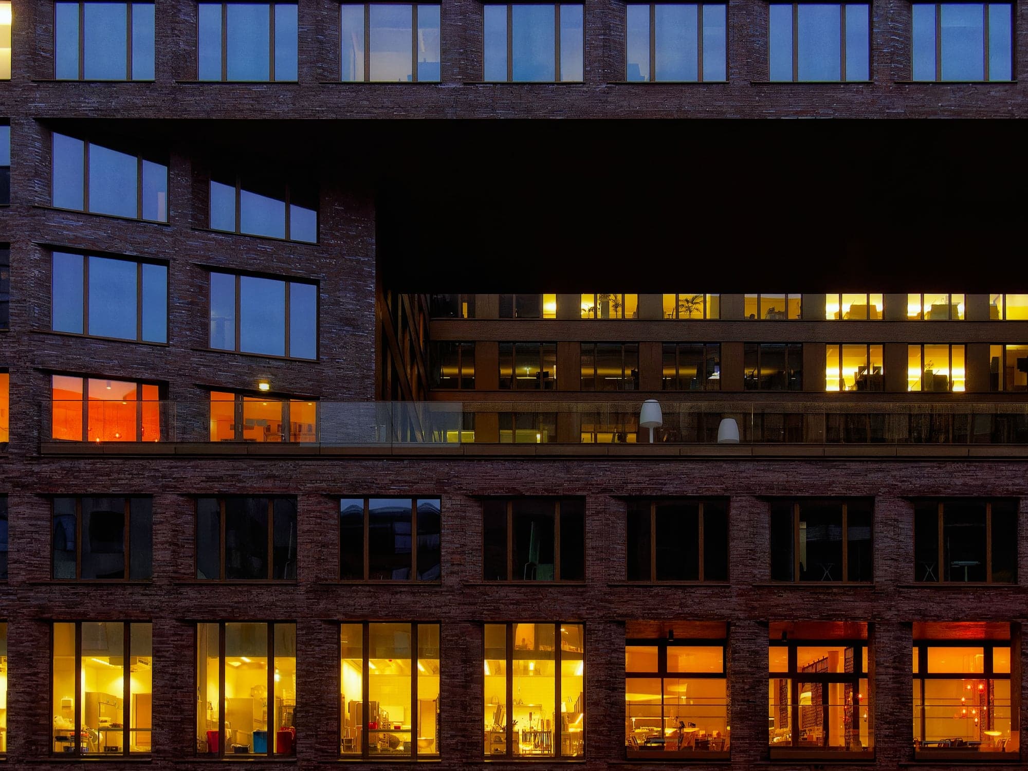 The windows of an office building at dusk, some of its rooms lit warm yellow.