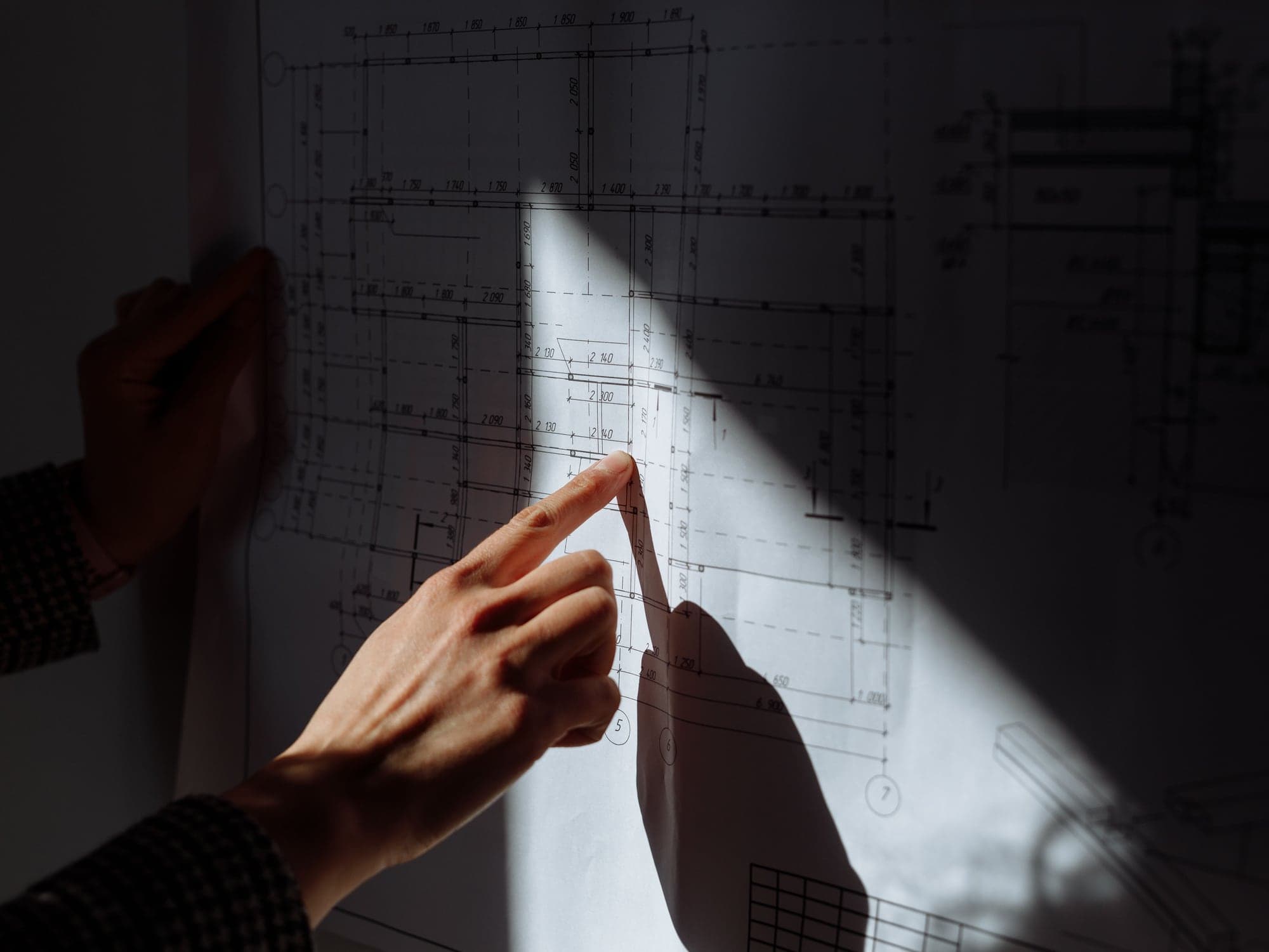 A hand pointing at the lines of a large drawing pinned to a wall, a beam of light across it.