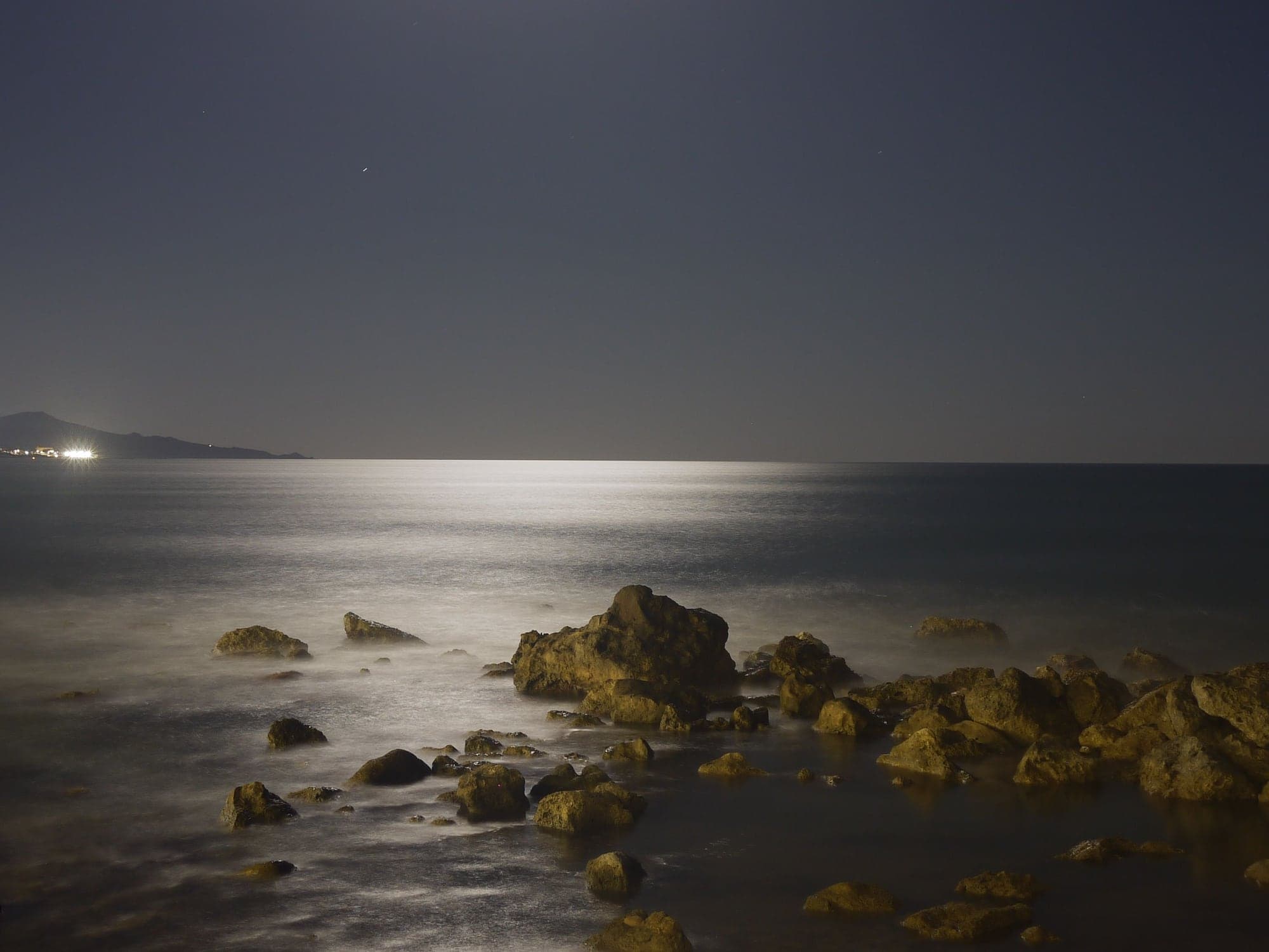 Moonlight on a calm sea at night, smooth water running around dark rocks.